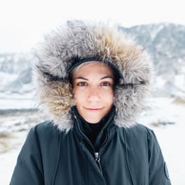 A smiling woman in a black coat with a furry hood stands outside in the snow, surrounded by mountains in the Lofoten Islands