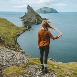 A girl in a red shirt stands on the coast of the Faroe Islands, with her hair blowing in the wind