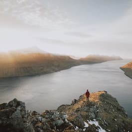 A girl stands on a rock outcropping in the Faroe Islands at sunrise, looking out onto the ocean