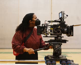 Woman behind a large camera wearing a black face mask and dark red sweater