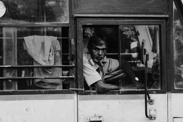 Black and white portrait of a man driving a vehicle, looking at the camera through an open window,