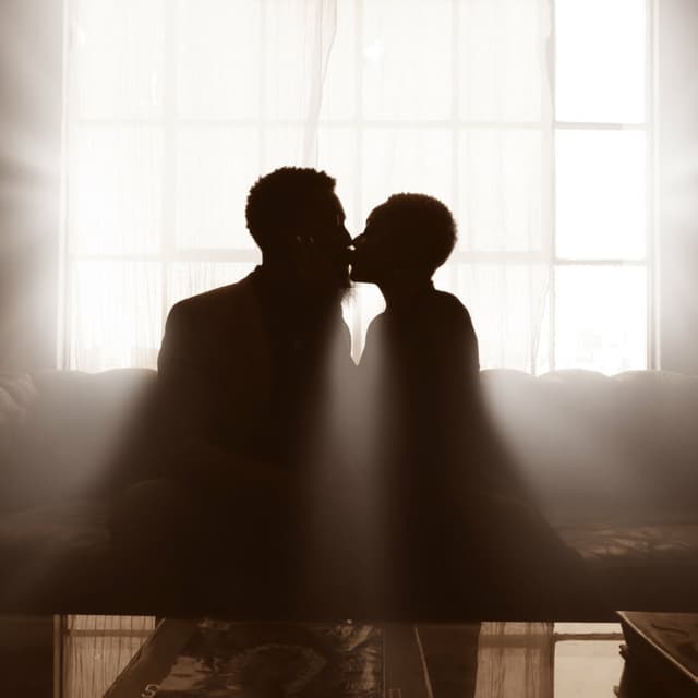 Two people sitting on a couch kissing with light from a window silhouetting them