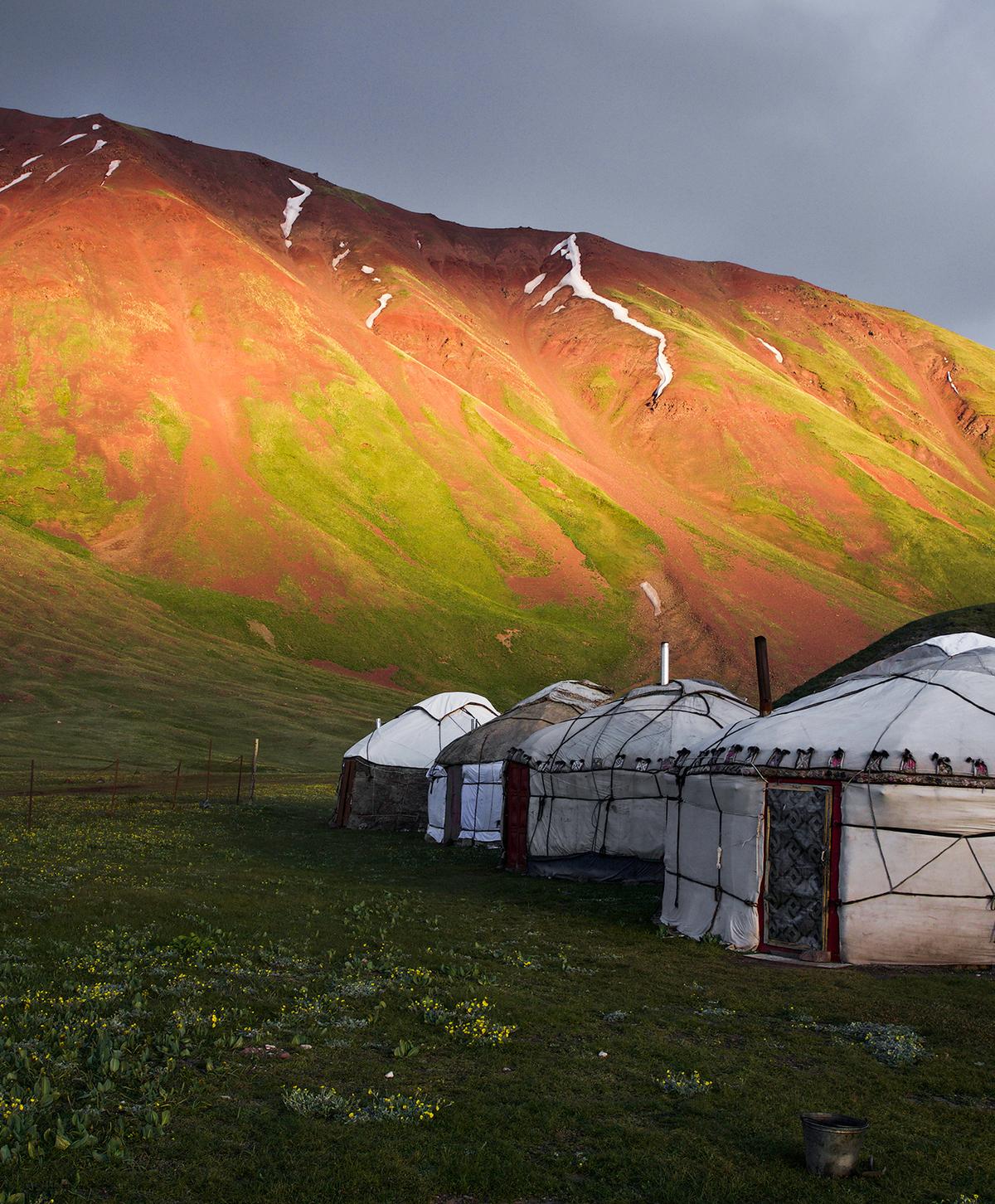 A yurt camp shines in the sunlight beneath an iconic red and green mountain in Kyrgyzstan