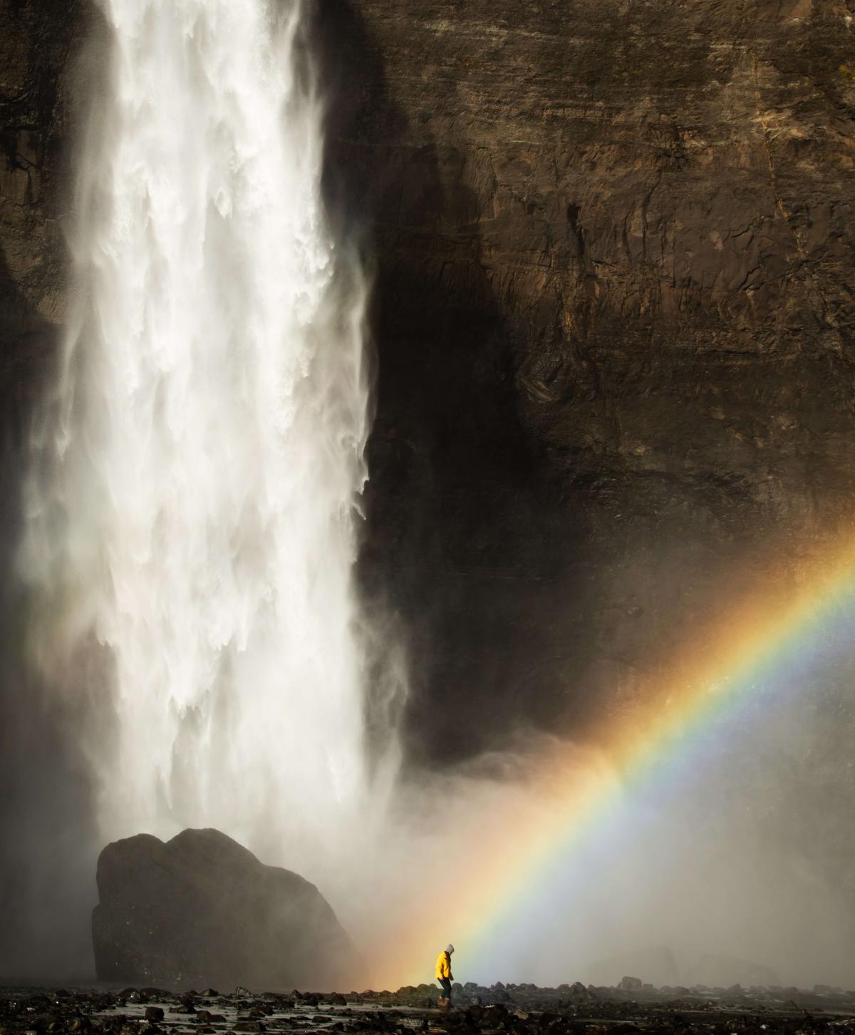 A girl in a yellow raincoat stands underneath rainbow at the bottom of a waterfall in Iceland