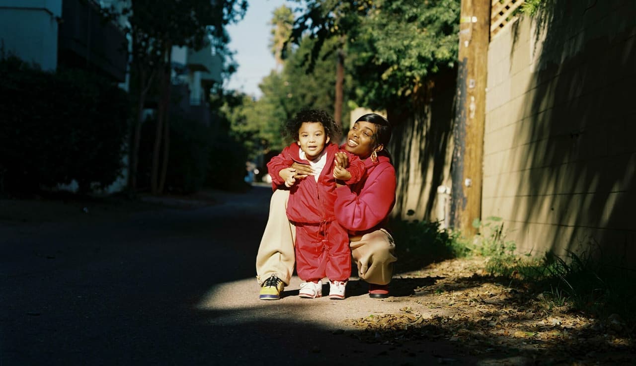 A woman smiling crouched near a child in a patch of sunlight on a street