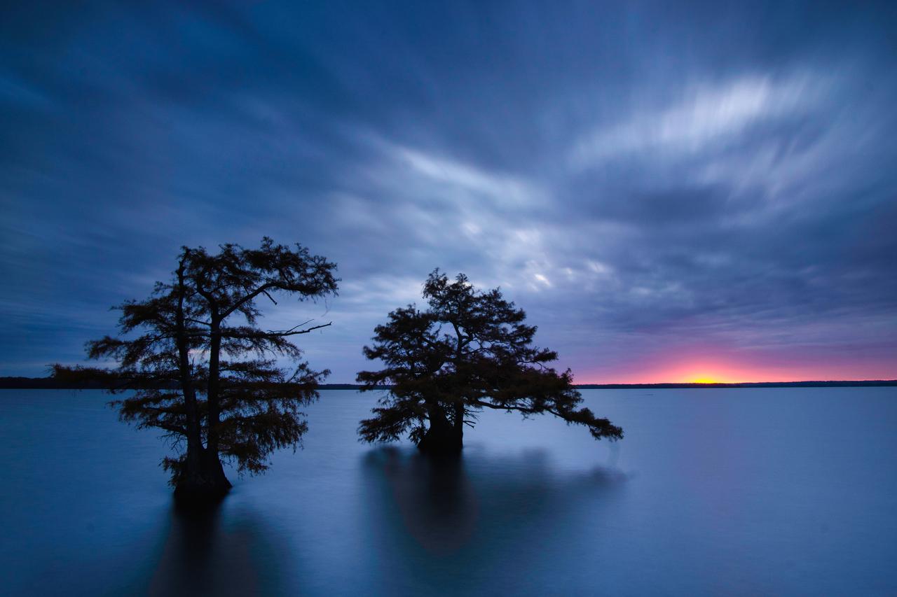 David Johnston - Long Exposure photo of clouds over a blue lake with two trees silhouetted