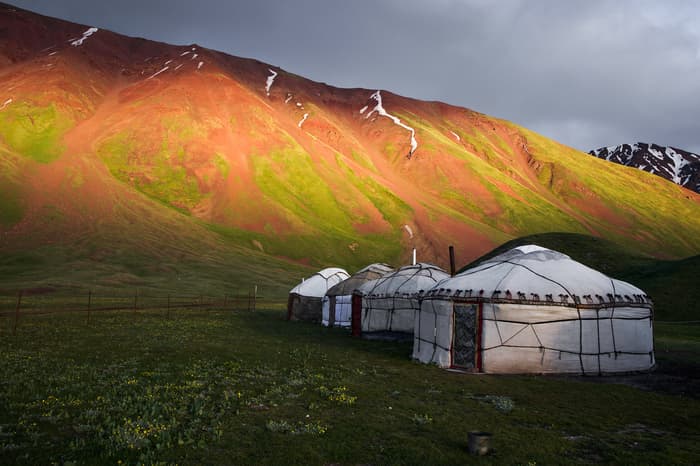 A yurt camp shines in the sunlight beneath an iconic red and green mountain in Kyrgyzstan